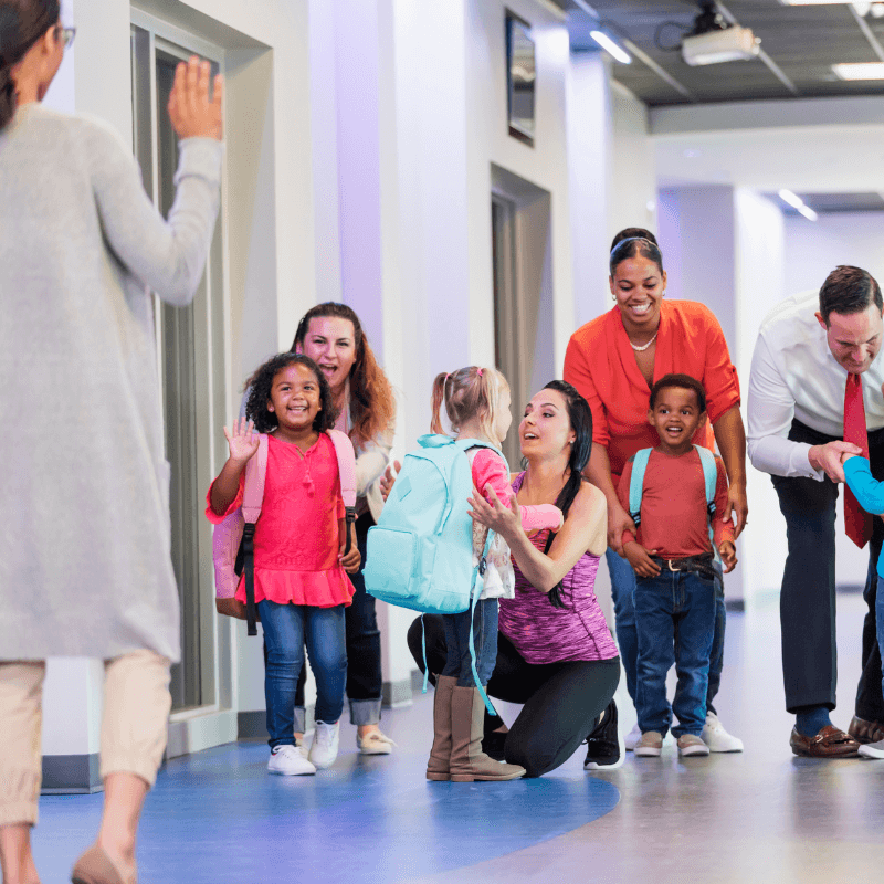 Teacher waves at parents as they drop of their students in the hallway. Strong connections and clear communication between teachers and school families greatly impacts school retention.