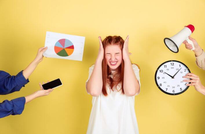 Caucasian woman overwhelmed by tasks holds hand up to ears and grimaces as people attempt to hand her various items: a chart, a phone, a clock, a megaphone. Each symbolizes a task or appointment. | Truth Tree partners with schools as their digital marketing partner. | Truth Tree provides digital marketing strategies and solutions for schools.