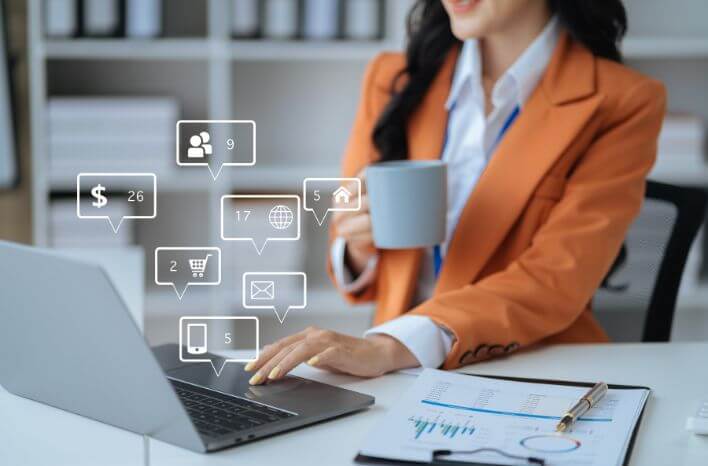 A woman holding a mug in one hand while the other sits atop a laptop keyboard with communication icons rising up from the lap top representing the different channels she has notifications on | this image accompanies a blog post by Truth Tree that focuses on strategic communication differentiating them from others because parents and students expect clear, concise, relevant communication.